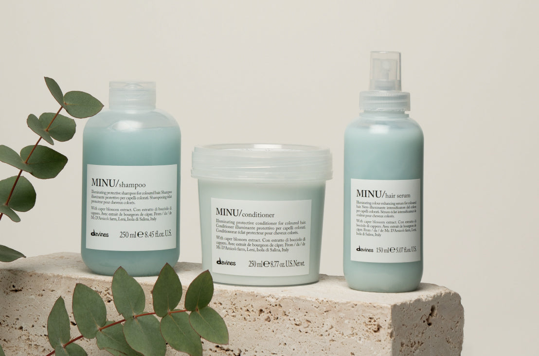 Shampoo, conditioner, and serum from Davines' color line, MINU, sit on a concrete block surrounded by green ferns